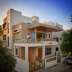 A daytime view of a modern family home, showcasing a cantilevered balcony with a wooden ceiling and glass railings. The design emphasizes clean lines and a connection between the upper living spaces and the outdoors.