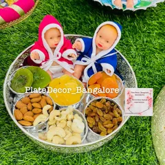A dry fruit platter for a baby shower celebrating twins. It features two baby dolls in pink and blue robes, surrounded by bowls of cashews, almonds, raisins, and other healthy snacks.
