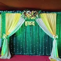 A simple and elegant Haldi backdrop for a home function. It features a green curtain with yellow and gold drapes, fairy lights, and a wooden "Haldi" sign.
