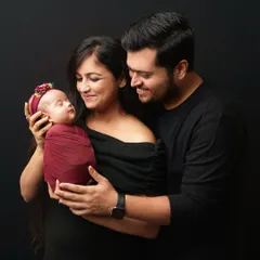 A beautiful family portrait where the mother holds the newborn while the father stands behind her, his arms gently wrapped around them both. Their coordinated black outfits create a cohesive and classic look.