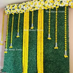 A classic Haldi or Mehendi backdrop using a green hedge wall as the base. The wall is decorated with vertical strings of yellow marigold flowers (genda phool) and white floral tassels. A thick border of yellow and white flowers across the top completes this traditional and fresh look.