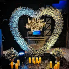 An elegant and dreamy proposal setup featuring a heart-shaped arch of pure white flowers. The glowing candles and central neon sign create a sophisticated and magical ambiance in a darker, intimate setting.