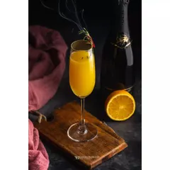 A classic mimosa, styled with a wisp of smoke from a rosemary sprig for a touch of drama. The dark background makes the bright orange juice stand out, while the Sola champagne bottle in the back provides context for a premium cocktail.