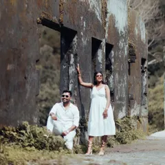 A stylish couple in matching white outfits poses against a rustic, weathered wall, blending modern fashion with a vintage-inspired location.