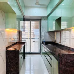 A full view of a sleek parallel kitchen layout in Ghansoli, Navi Mumbai. The high-gloss mint green cabinets reflect light beautifully, making the space feel open and bright, while the dark granite countertops provide a durable work surface.