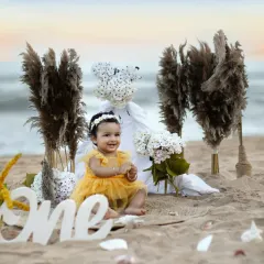 A happy baby girl claps her hands with excitement during her "One" birthday photoshoot on the sand, surrounded by boho-style props.