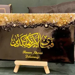 This personalized plaque features the verse "Fabi ayyi ala i rabbikuma tukazziban" in bold yellow calligraphy on a black background. The top is adorned with a textured mix of gold and silver embellishments, and it includes the client's name for a personal touch.