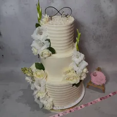 A timeless two-tier wedding cake with a classic ridged buttercream finish. It's adorned with fresh white gladiolus and roses, and topped with an infinity 'Love' sign, perfect for an elegant and traditional celebration.