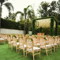 An outdoor ceremony seating arrangement with elegant wooden chairs on a lush green lawn. The aisle is clear, leading to a beautiful mandap covered in greenery.