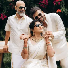 A modern family portrait, captured outdoors with natural light. The son embraces his mother, who is seated, while the father stands beside them, all sharing a moment of relaxed connection in coordinated white outfits.