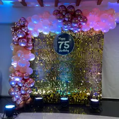 A glamorous 75th birthday photo backdrop featuring a gold shimmer wall, a custom "Happy 75 Birthday" sign, and an elegant balloon garland, all highlighted by professional uplighting.