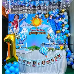 For this first birthday, I designed a sky and hot-air balloon theme. The decor features a custom backdrop, blue and white balloon clouds, a photo banner showing the baby's first year, and a large '1' foil balloon.