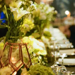 A close-up of a tablescape, where a geometric candle holder casts a warm glow on a lush arrangement of white and green florals.