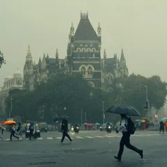 Pedestrians cross a wet street with the historic High Court building looming in the background. The image captures the daily rhythm of Mumbai life that continues, beautifully and stoically, even in the middle of a downpour.