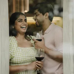 A playful, candid moment from a pre-wedding shoot, viewed through a window. The couple's shared laughter over a glass of wine creates an intimate and authentic portrait.