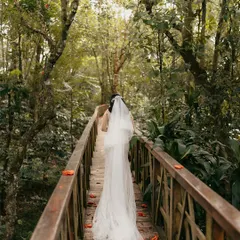 A bride walks alone across a wooden bridge deep in a lush forest. The long veil trailing behind her and the canopy of trees create a serene and magical atmosphere, perfect for a nature-inspired wedding.
