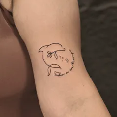 A playful and simple single-line dolphin tattoo. The design incorporates a flower and sparkling stars, with a delicate script that reads "Todo a su tiempo" (Everything in its own time).