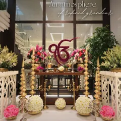 The heart of the ceremony setup, featuring a floral Ek Onkar symbol crafted from deep red carnations. This sacred centerpiece is surrounded by lush flower arrangements, traditional brass lamps, and elegant white furniture to create an atmosphere of refined grandeur.