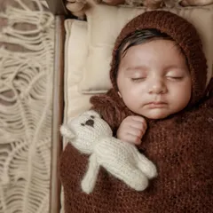 A close-up portrait of the baby in the brown swaddle and bonnet, highlighting their delicate features. The earthy color palette is both modern and timeless.