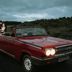 The couple shares an embrace in their vintage car as dusk settles, with the headlights on. This creates a moody, cinematic, and incredibly romantic atmosphere.