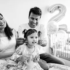 A candid black and white family photo from a second birthday shoot. The parents are sharing a laugh while their daughter looks on, capturing a genuine slice of their family life.
