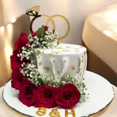 A cake to celebrate a promise. This engagement cake is adorned with a cascade of real red roses, baby's breath, and a topper of two gold rings, symbolizing love and commitment.