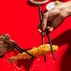 A dynamic shot of two people interacting with crispy shrimp tempura against a bold red background for Harajuku Tokyo Cafe.