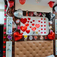 A wall decorated for a romantic surprise, featuring numerous red heart cutouts, heart-shaped foil balloons, and cute couple-themed stickers.
