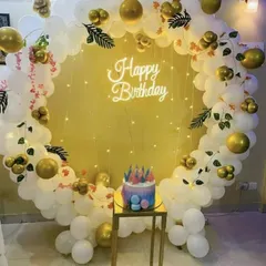 An elegant white and gold balloon ring, perfect for cake cutting. The setup includes a 'Happy Birthday' neon sign, fairy lights, and a gold pedestal for the cake.