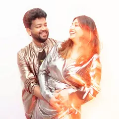 A joyful, candid moment captured with a warm light flare effect. The couple laughs together, their shiny outfits catching the light and adding to the photo's dynamic, celebratory energy.