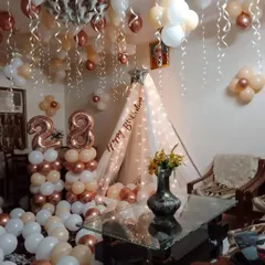 A cozy and stylish 28th birthday room decoration. This features a canopy tent with fairy lights, rose gold and white balloons covering the floor and ceiling, and large foil number balloons.