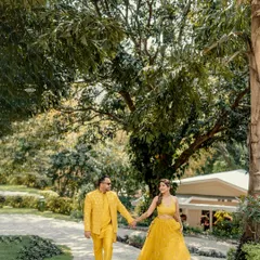 A happy stroll through a sunlit garden path. The vibrant yellow outfits pop against the green scenery, and their hand-in-hand walk creates a feeling of easy, comfortable love. This is a perfect shot for a Mehendi or Haldi themed pre-wedding session.