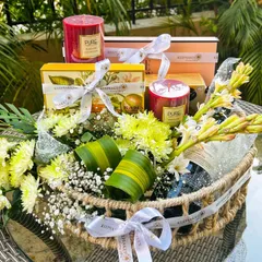 This elegant hamper was customized for a bride and groom, matching their wedding's white and green color scheme. It features fragrant candles, a bottle of sparkling wine, and other treats nestled in a jute basket with fresh chrysanthemums and tuberoses.