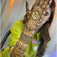 A bride peeking through her stunning back-of-hand mehndi. The design is personalized with the couple's initials "DV" and "S", surrounded by intricate traditional patterns.
