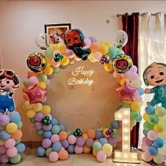 This Cocomelon first birthday party features a full ring of pastel balloons, character cutouts on balloon pillars, a 'Happy Birthday' neon sign, and a light-up number '1'.