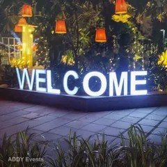 A bold, illuminated "WELCOME" sign makes a clear and stylish statement at the entrance of any event. Surrounded by lush greenery and hanging lanterns, it creates an inviting and festive atmosphere for arriving guests.