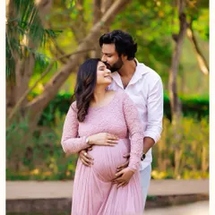 A tender outdoor portrait where a partner embraces the mother-to-be from behind, both looking down at her baby bump with love. The soft natural light filters through the trees, highlighting this sweet moment.