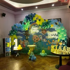 A grand Cocomelon first birthday party setup in a hall, featuring a full backdrop, a vibrant balloon arch, two neon "Happy Birthday" signs, and marquee letters for the child's name.