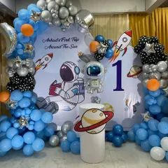 Let's get creative with a unique theme for your little one's first birthday. This 'First Trip Around The Sun' space-themed party featured a custom backdrop with astronauts and rockets, surrounded by a galaxy of blue, silver, and black balloons.