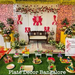 An outdoor seer varisai setup with a traditional backdrop. The display includes a variety of platters, from decorated fruits to doll arrangements, all set on a lush green base.