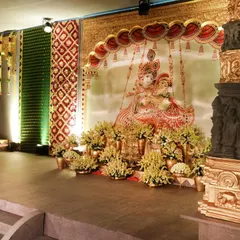 A side stage within our temple-themed wedding, featuring a large Tanjore-style painting of Radha and Krishna. The area is flanked by intricately carved pillars and adorned with yellow flowers, serving as a beautiful photo backdrop and lounge space.