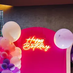 A vibrant "Happy Birthday" neon sign in a warm orange glow, set against a bold magenta backdrop and surrounded by balloons.