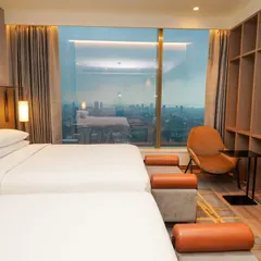 A hotel-style bedroom with two beds, showcasing the clean lines of the furniture, built-in shelving, and a large window with a cityscape backdrop.