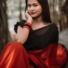 A classic seated pose in a beautiful red and black saree. The natural forest-like background adds a rustic feel to this ethnic portrait, highlighting the simple elegance of the attire.