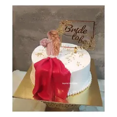 A chic and stylish "Bride to be" cake. The design includes a beautiful illustration of the bride, a flowing red fondant dress, and a custom gold topper, all on a base of elegant white buttercream.