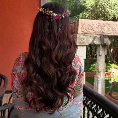 A beautiful half-up hairstyle for a Haldi ceremony. The hair is styled in soft waves and crowned with a halo of colorful, fresh flowers, creating a bohemian and festive look.