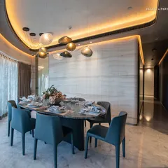 The dining area at Lodha World View follows the apartment's signature curved architecture. A bespoke oval dining table is surrounded by elegant blue chairs, all under a cluster of hand-blown glass pendant lights that add a touch of fluid artistry.