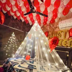 A cozy and romantic room decoration featuring a tent made of sheer fabric and fairy lights over the bed. The ceiling is filled with red and white balloons, and 'Just Married' foil letters are placed on the headboard.