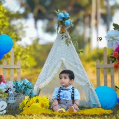 A baby boy sits in front of a teepee at a private outdoor location. The natural greenery and soft sunlight create a beautiful and peaceful setting for his portraits.