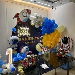 This space-themed setup for Sanidhya's first birthday was designed to fit perfectly in a home setting. It features a rocket ship cutout, a "Happy Birthday" neon sign, and astronaut foil balloons.